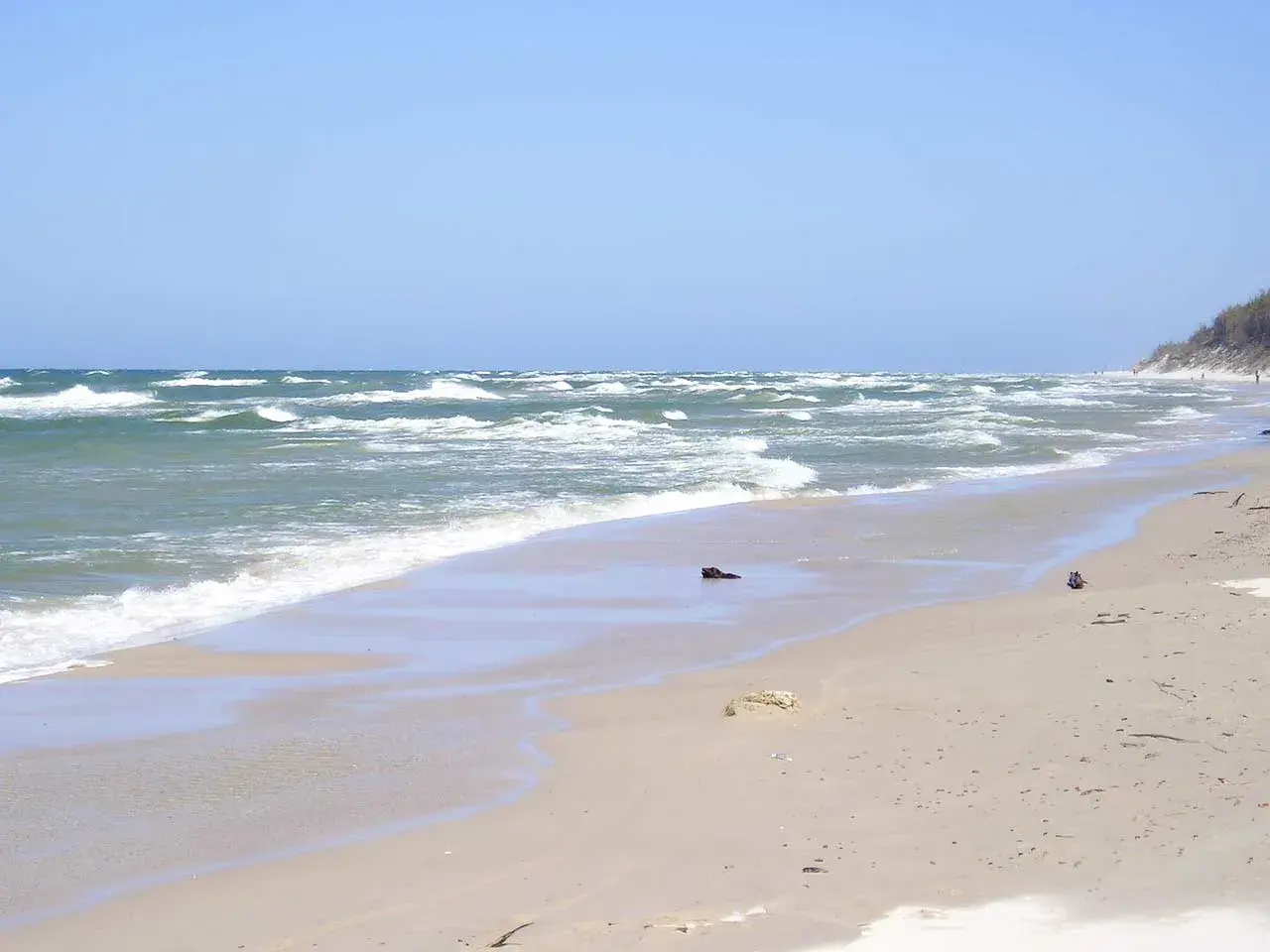 Plaża w Rowach, z falującym morzem i piaszczystym brzegiem. W oddali widać wydmy i nielicznych spacerowiczów.