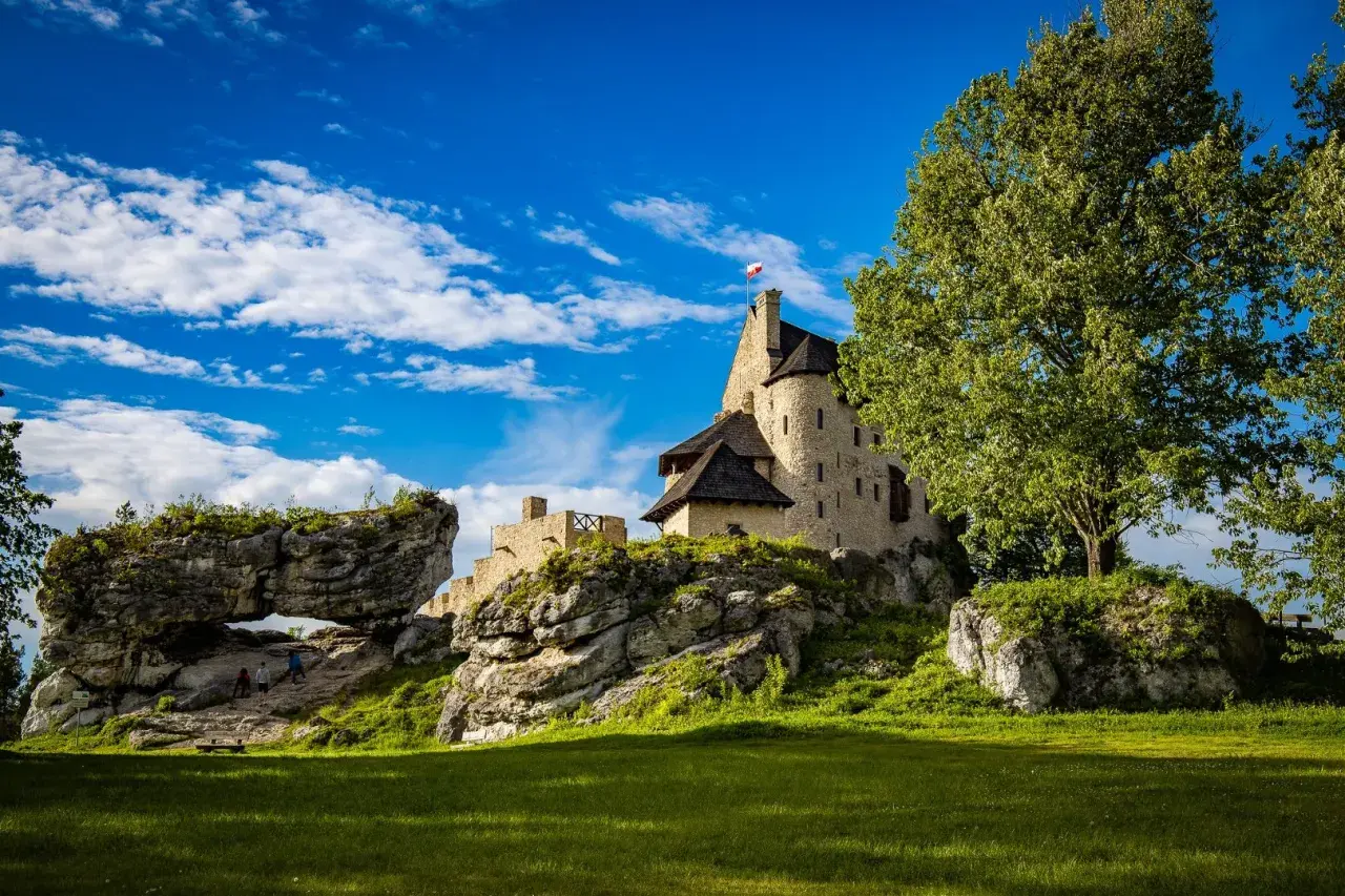 Kamienny zamek na skale, z polską flagą, otoczony zielenią i skalnymi formacjami. W oddali widać łuk skalny, przez który przechodzą turyści. To malownicze mirów bobolice.