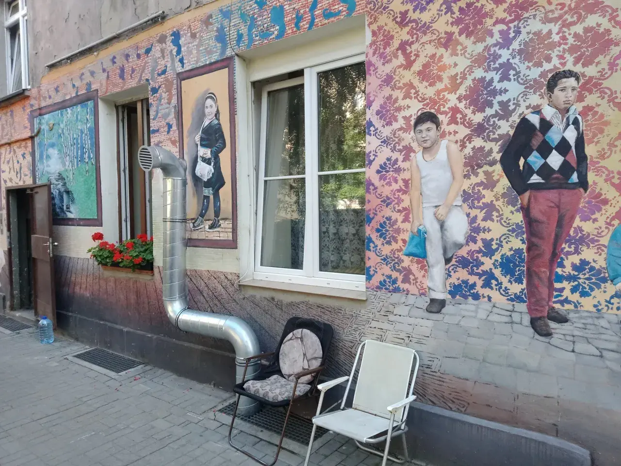 Kolorowe murale z postaciami dzieci i wzorami zdobią kamienicę na Nadodrzu. Dwa krzesła stoją przed ścianą.
