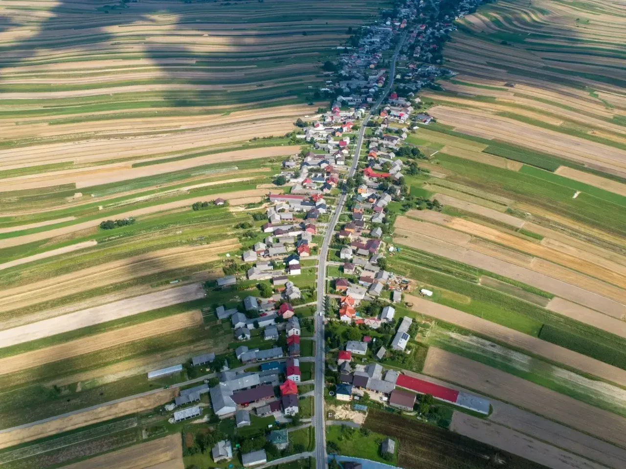 Widok z lotu ptaka na długą wieś w Polsce, rozciągającą się wzdłuż drogi, otoczoną polami o różnych kolorach.