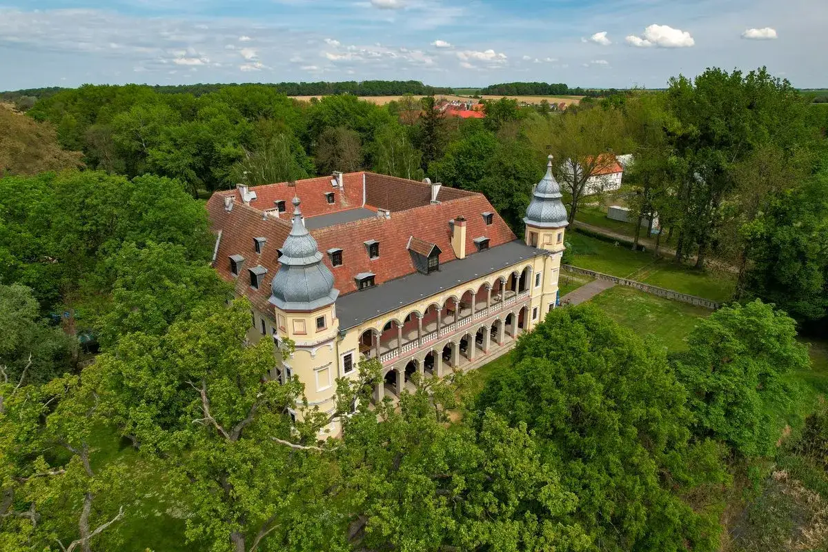 Pałac w Nowej Wsi Wrocławskiej otoczony zielenią, z charakterystycznymi wieżyczkami i arkadowym krużgankiem.