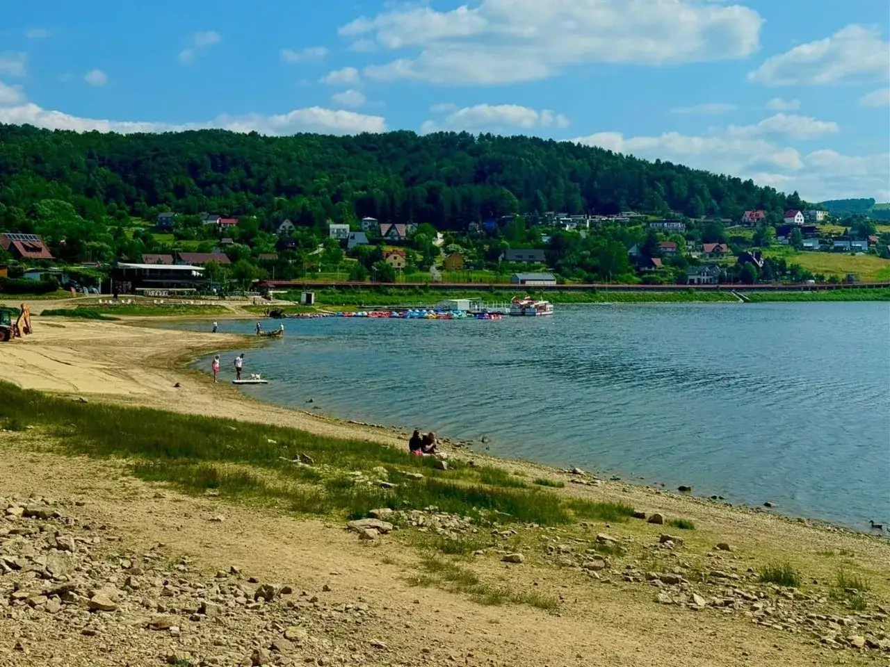 Jezioro Rożnowskie oferuje liczne atrakcje: piaszczysta plaża, wypożyczalnia sprzętu wodnego, a w tle malownicze wzgórza z domkami.