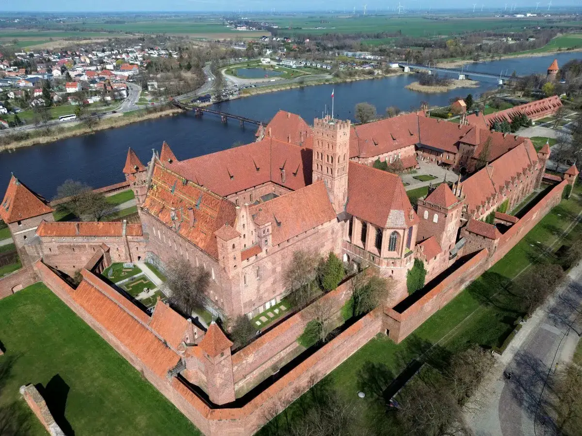 Potężny zamek krzyżacki z czerwonej cegły, otoczony murami obronnymi, nad rzeką. W tle miasto i pola.