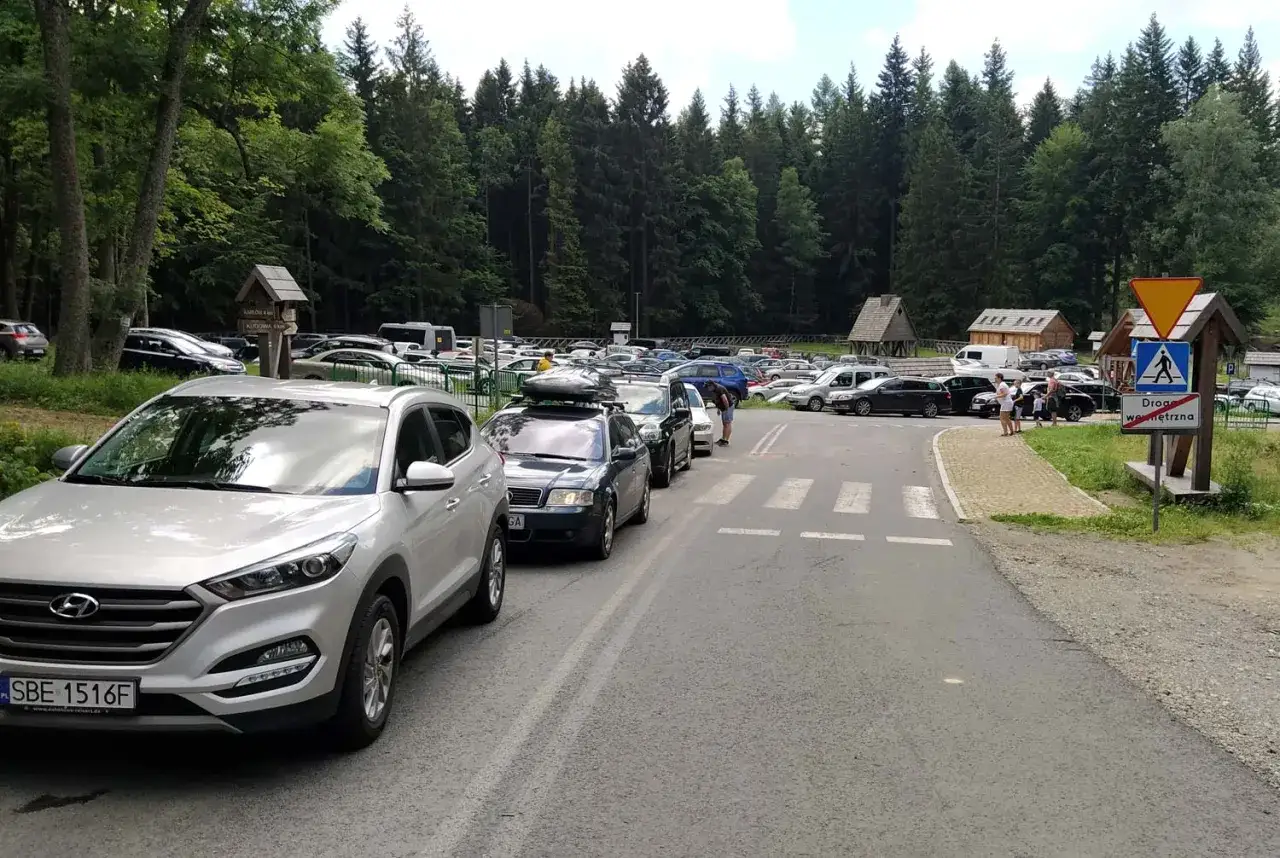 Parking przy Błędnych Skałach, pełen samochodów. W oddali widać budynki i las. Brak widocznego cennika.