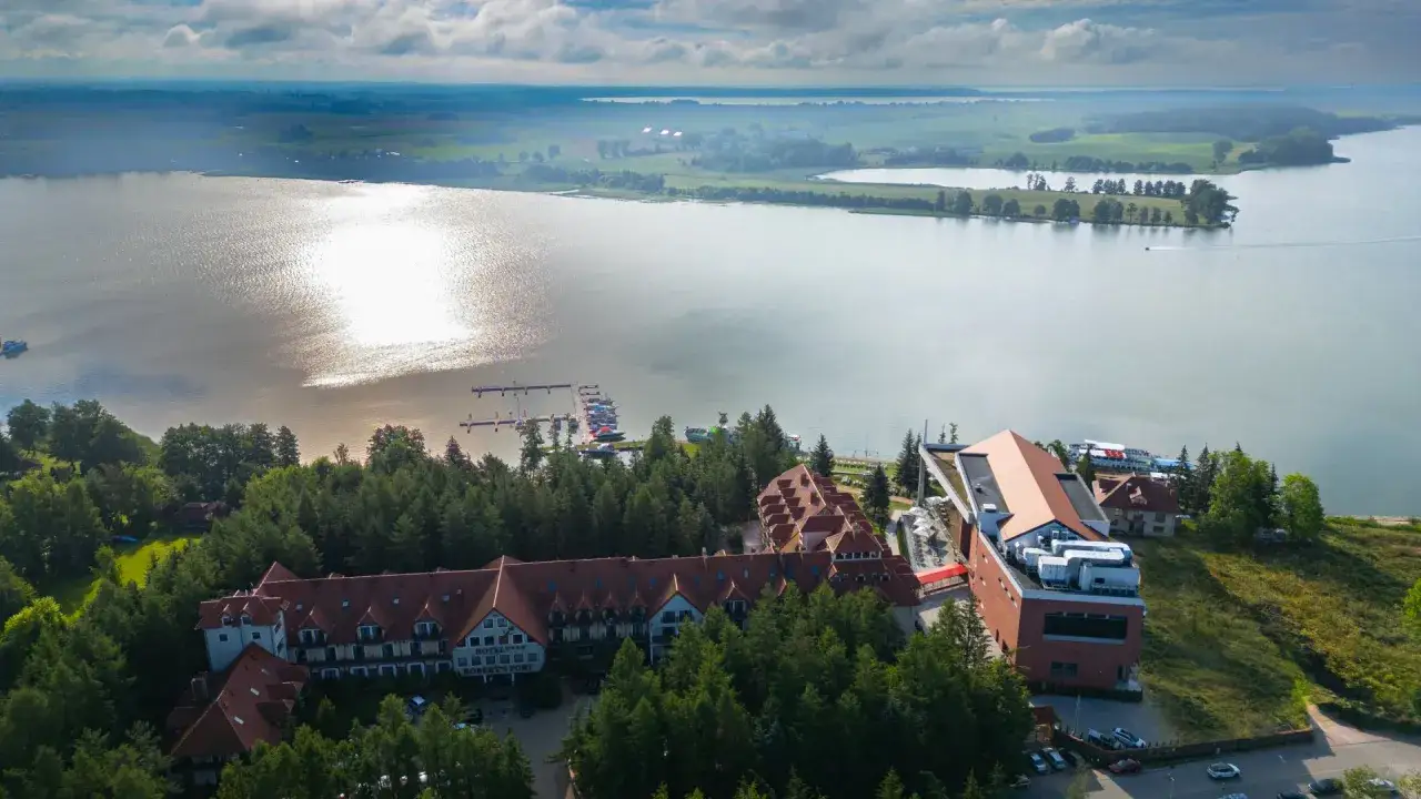 Hotel Mikołajki Leisure & Spa z lotu ptaka. Widok na jezioro, przystań z łodziami i hotel otoczony lasem.