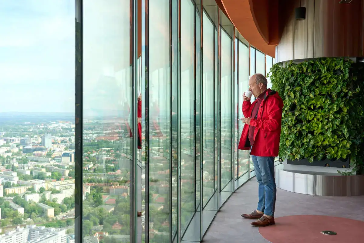 Mężczyzna w czerwonej kurtce pije kawę, patrząc na panoramę Wrocławia z Sky Tower. Sprawdź Sky Tower Wrocław cennik, by poznać ceny biletów.