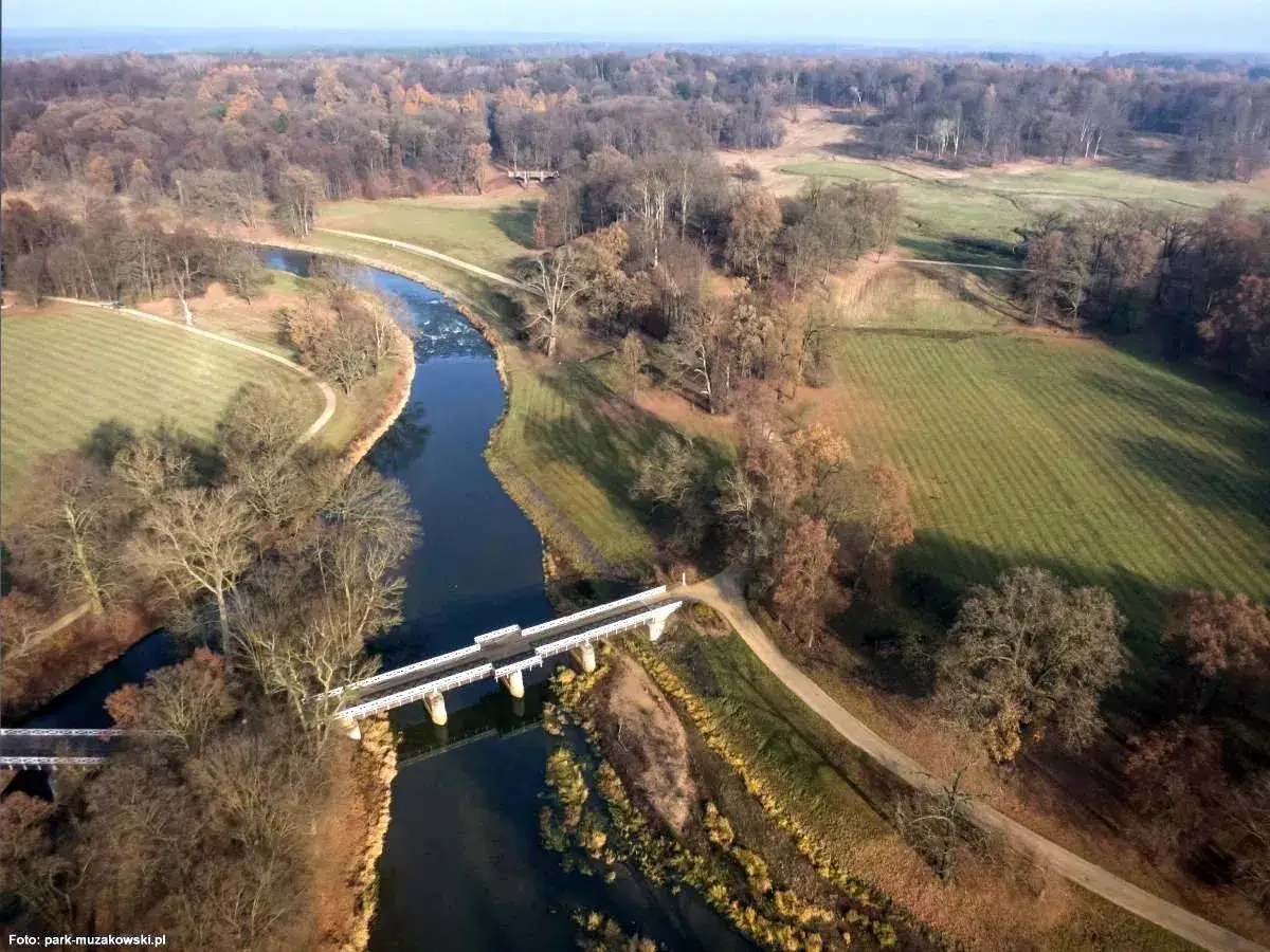 Malowniczy park mużakowski zachęca do zwiedzania. Rzeka przepływa przez krajobraz z mostami i alejkami wśród jesiennych drzew.