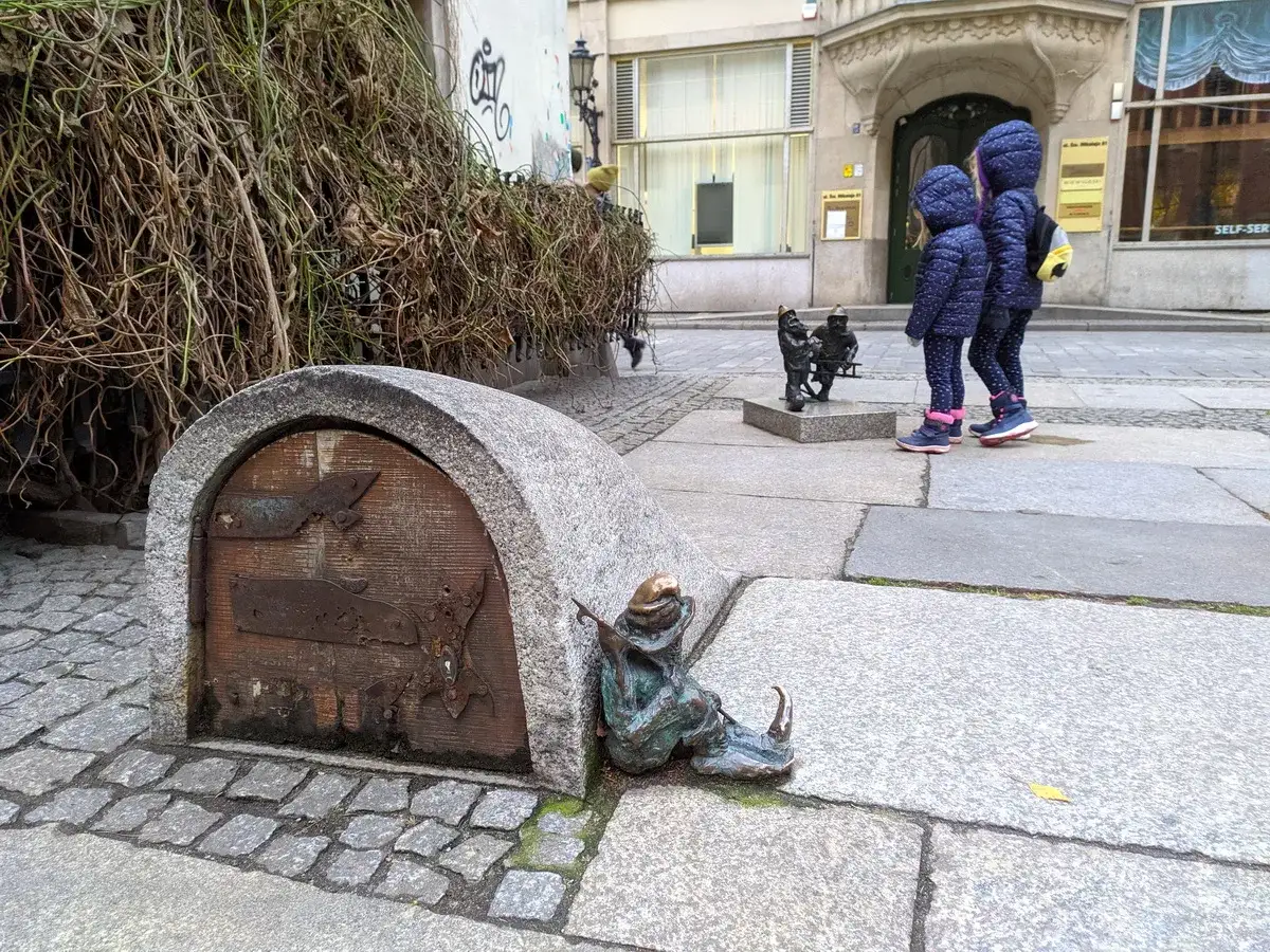 Dzieci obserwują krasnale we Wrocławiu. Ile krasnali jest we Wrocławiu? Trudno policzyć wszystkie, ale ten siedzący przy kamiennym domku jest uroczy.