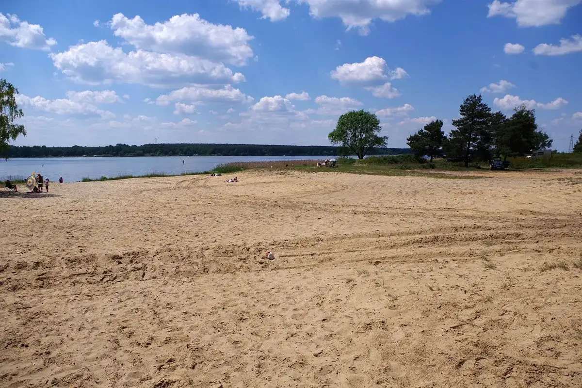 Piękny, piaszczysty poraj nad jeziorem. Kilka osób odpoczywa na plaży, ciesząc się słońcem i spokojem.
