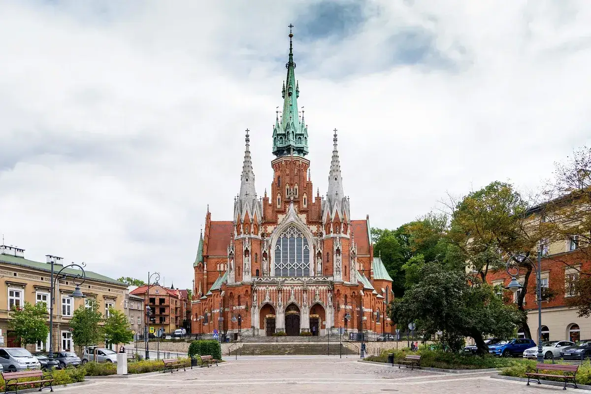 Imponujący kościół na rynku podgórskim, z ceglaną fasadą i zielonymi wieżami, otoczony drzewami i budynkami.