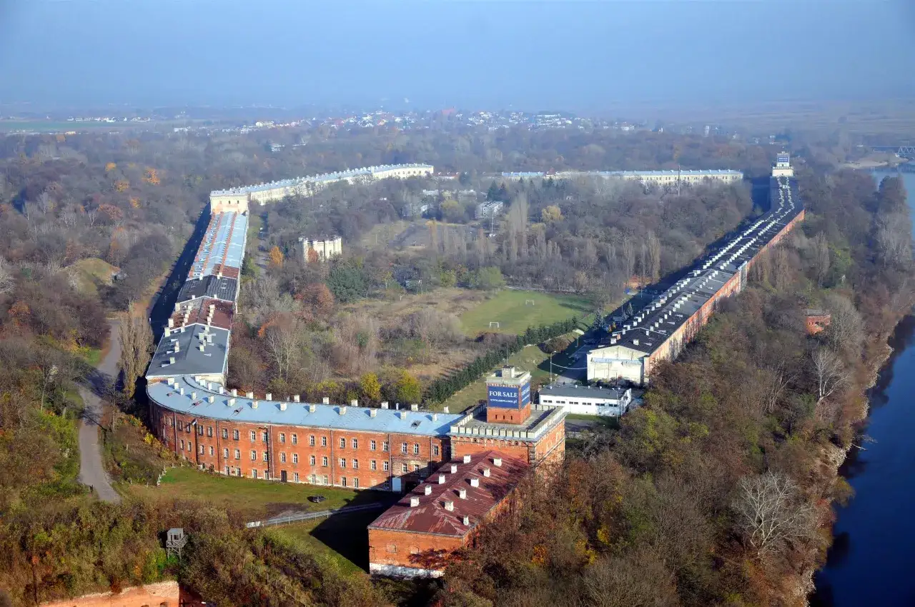 Widok z lotu ptaka na Twierdzę Modlin, idealne miejsce na zwiedzanie. Długie, ceglane budynki otoczone drzewami.