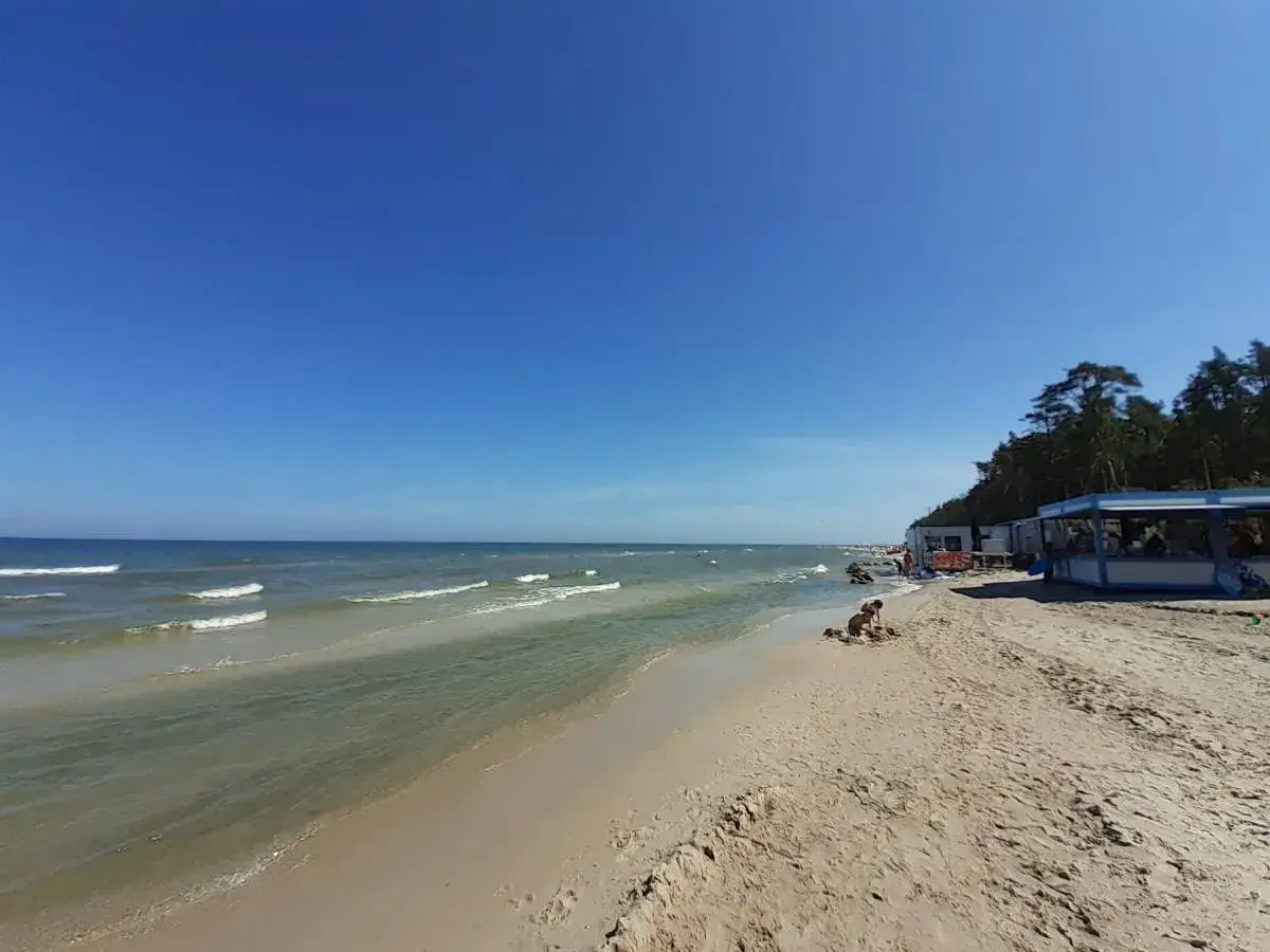 Słoneczna lubiatowo plaża z łagodnymi falami i budynkami w tle. Dziecko bawi się piaskiem.
