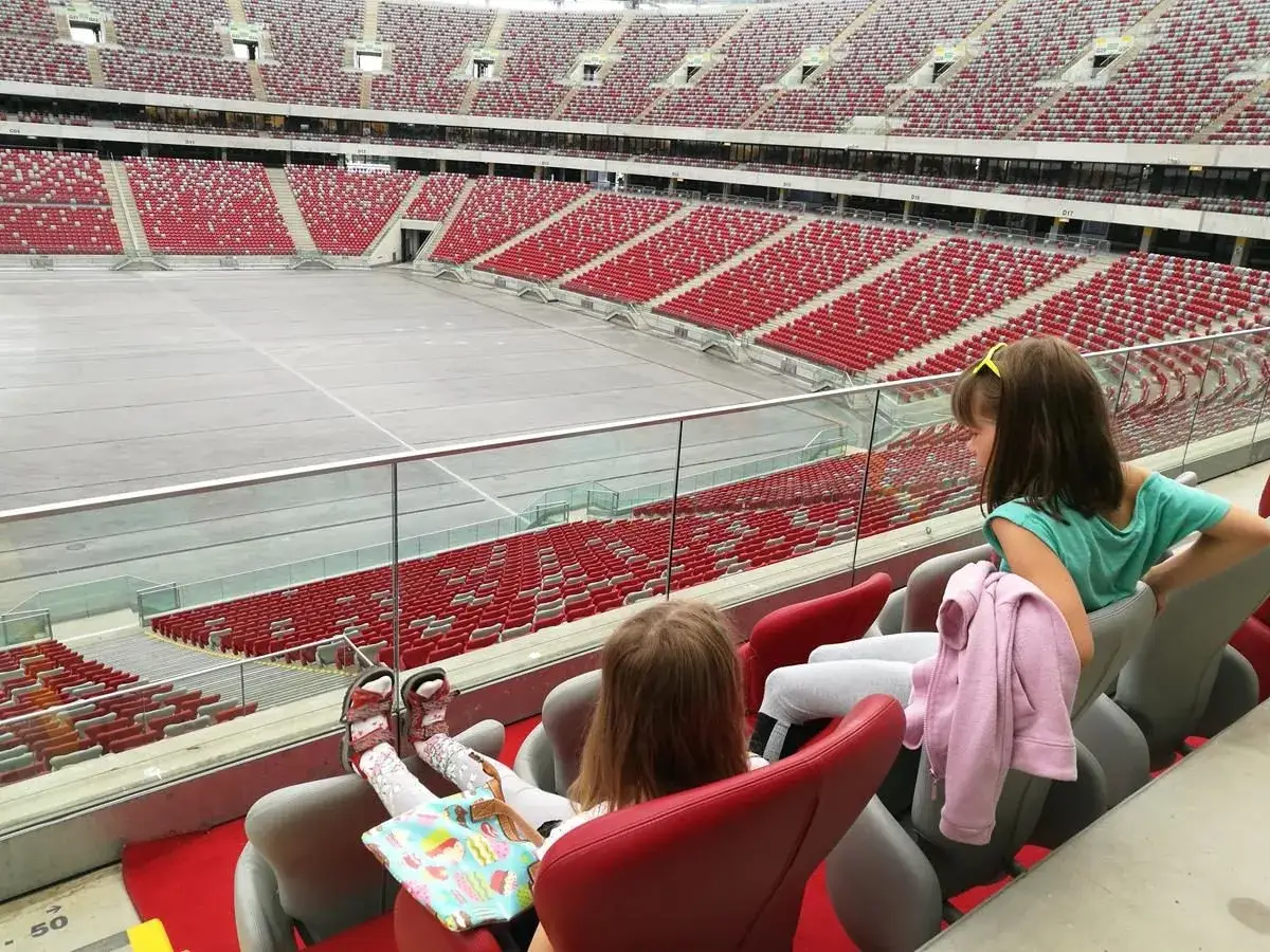 Dwie dziewczynki siedzą na trybunach stadionu, podziwiając puste boisko. Zastanawiają się, ile miejsc ma stadion narodowy.