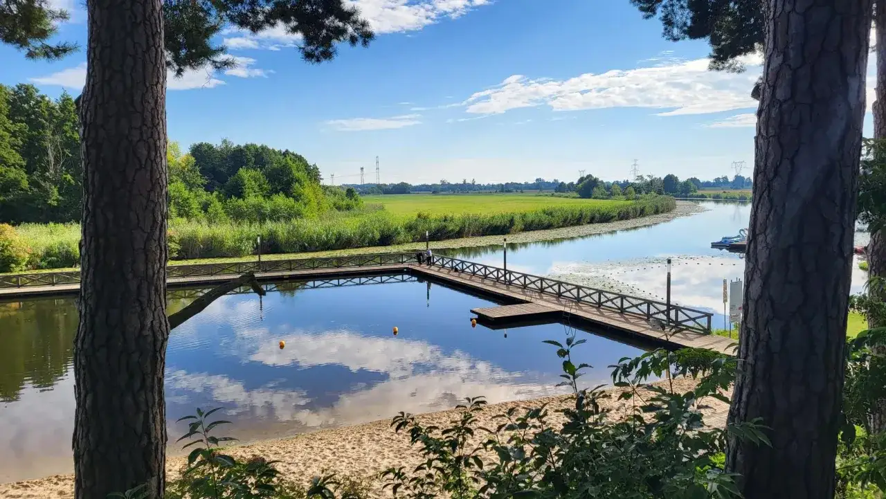 Spokojne jezioro Kozienickie z drewnianym pomostem i piaszczystą plażą, otoczone zielenią i błękitnym niebem z chmurami.
