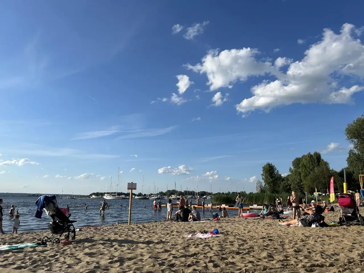 Słoneczna lubczyna plaża tętni życiem. Ludzie kąpią się, opalają i odpoczywają na piasku, podczas gdy na wodzie kołyszą się żaglówki.