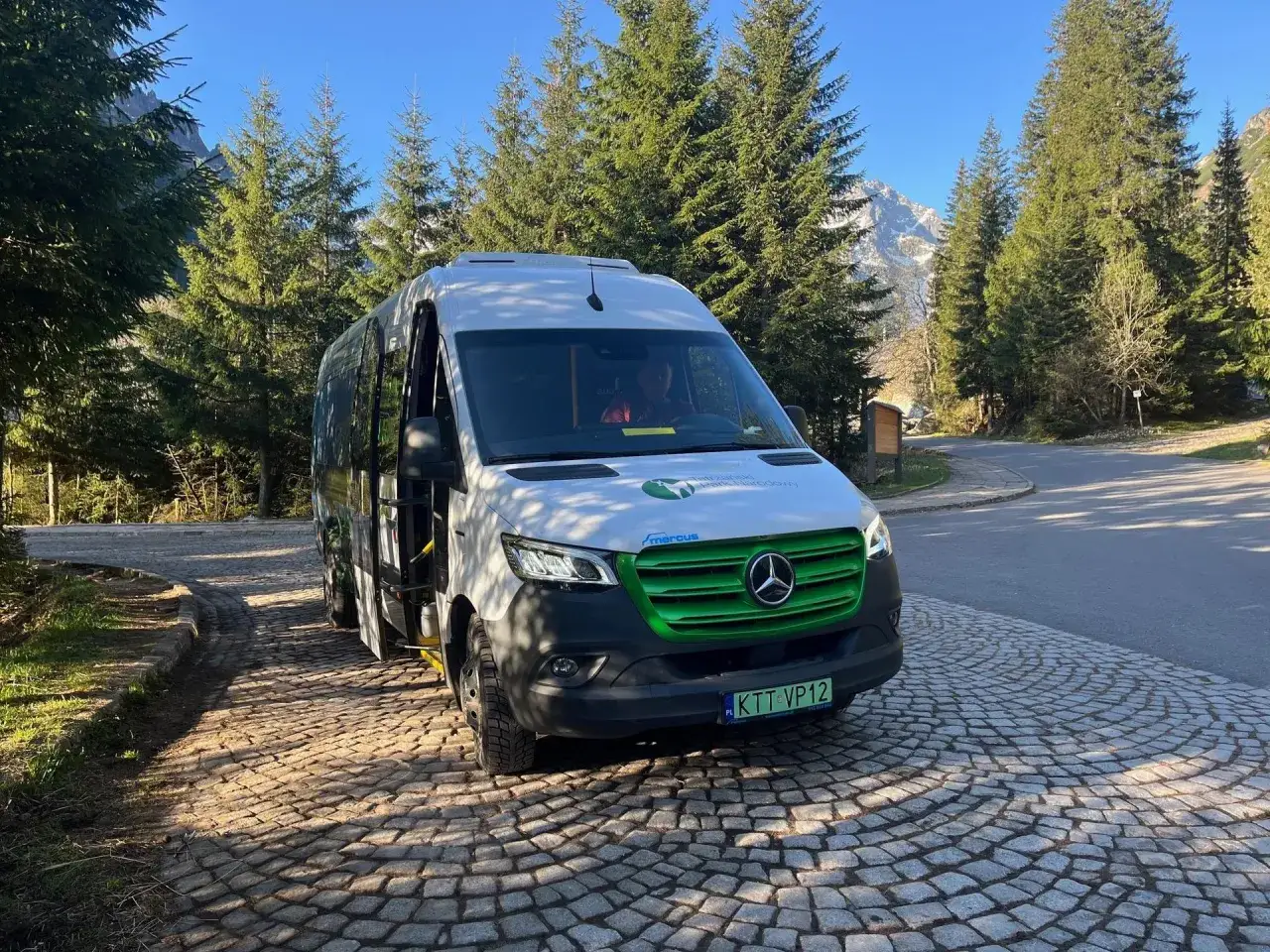 Busy na Morskie Oko czekają na turystów. Biały bus z zielonym grillem stoi na brukowanej drodze wśród drzew, w tle góry.